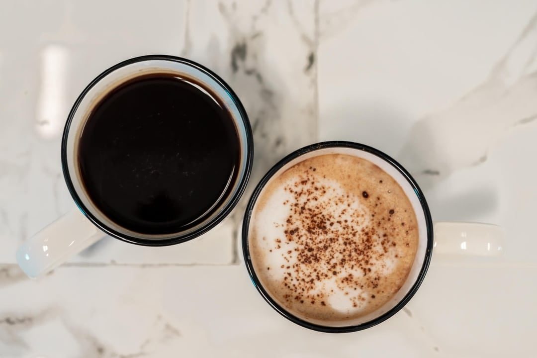 Overhead view of a black coffee and a cappuccino in enamel mugs on marble